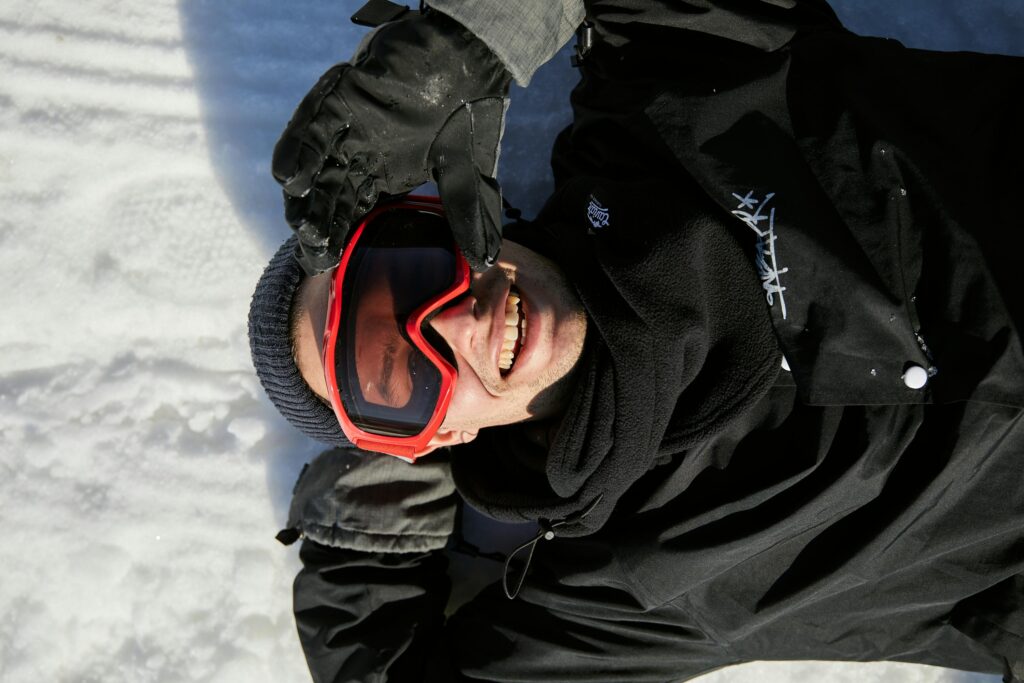A joyful man wearing protective goggles smiles while lying on snow in winter attire.